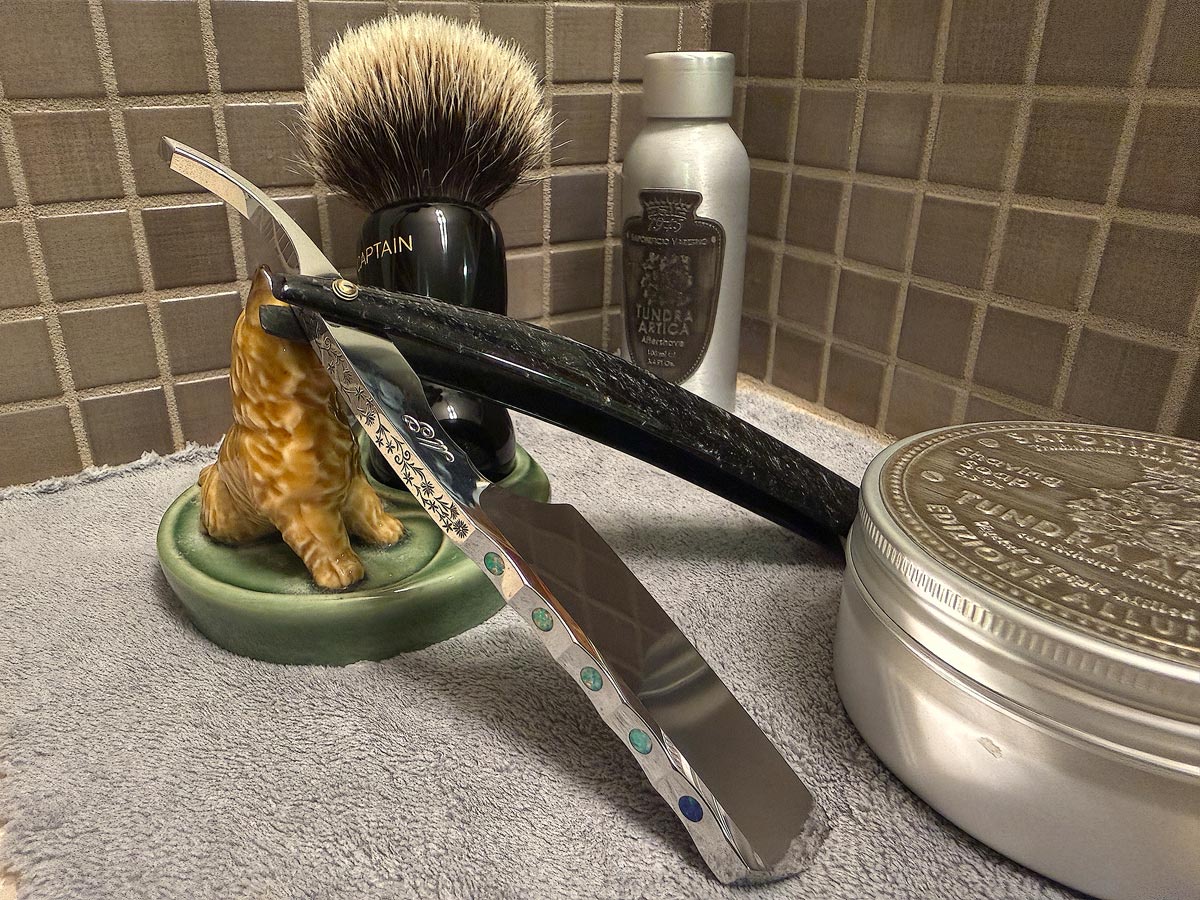 On a gray microfiber towel a straight razor with blade open about 45 degrees leans next to a bottle of aftershave and a jar of shave soap. A shave brush rests bristles-up in a green Wade ceramic pipe rest that features a brown Cairn Terrier.