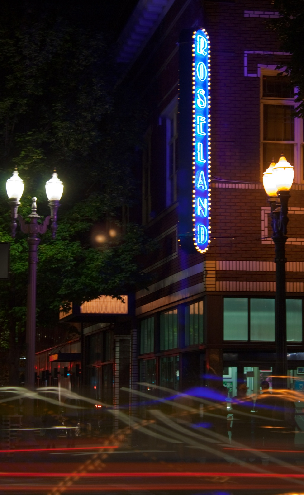 the roseland theater neon sign in portland, or, from across the intersection. there are light trail and reflections from the traffic, and an elegant streetlamp tries its best to illuminate, but the blue of the neon stands out against the dark brick facade. 