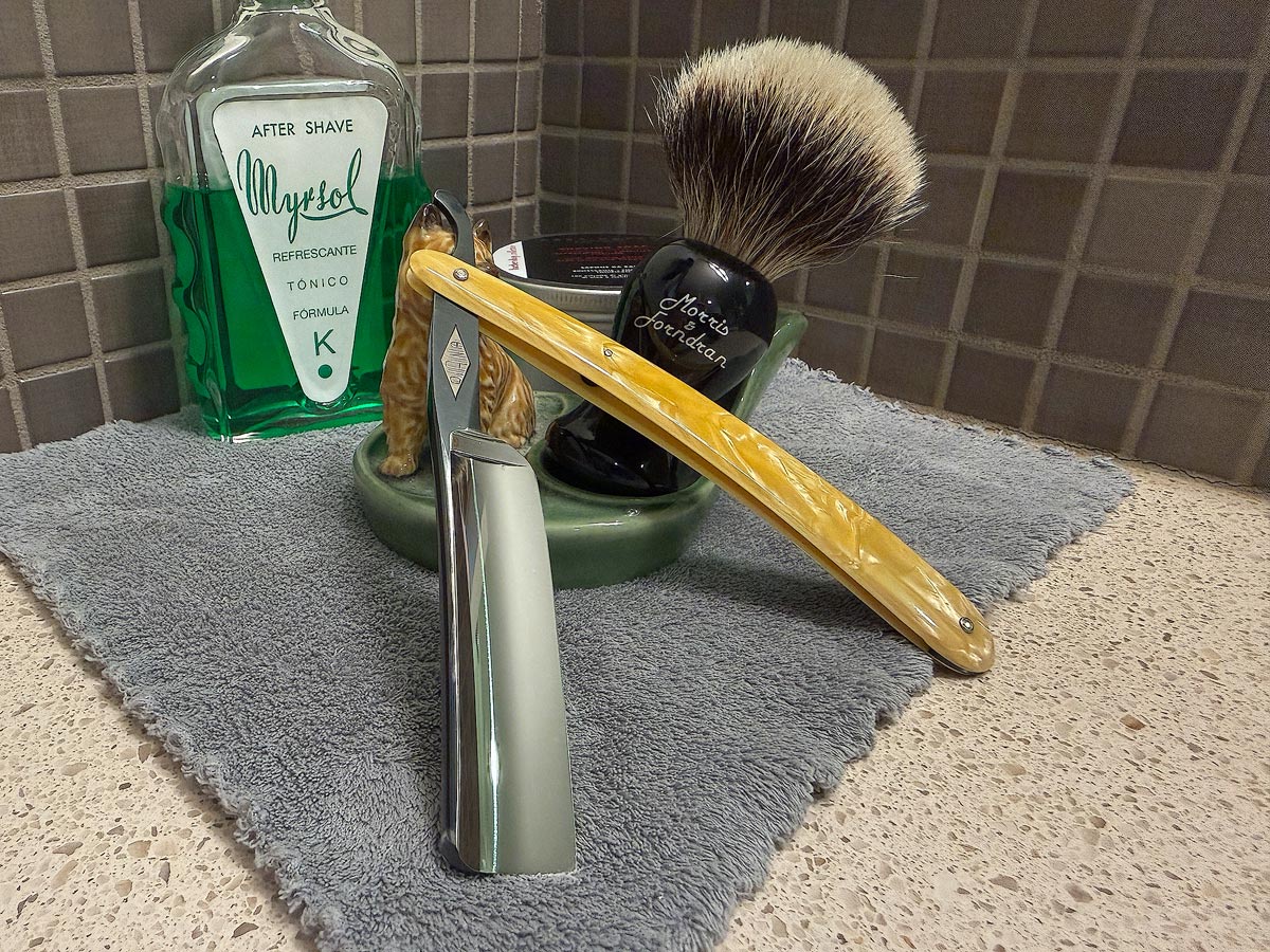 On a gray microfiber towel a straight razor with blade open about 45 degrees leans next to a bottle of aftershave and a jar of shave soap. A shave brush rests bristles-up in a green Wade ceramic pipe rest that features a brown Cairn Terrier.