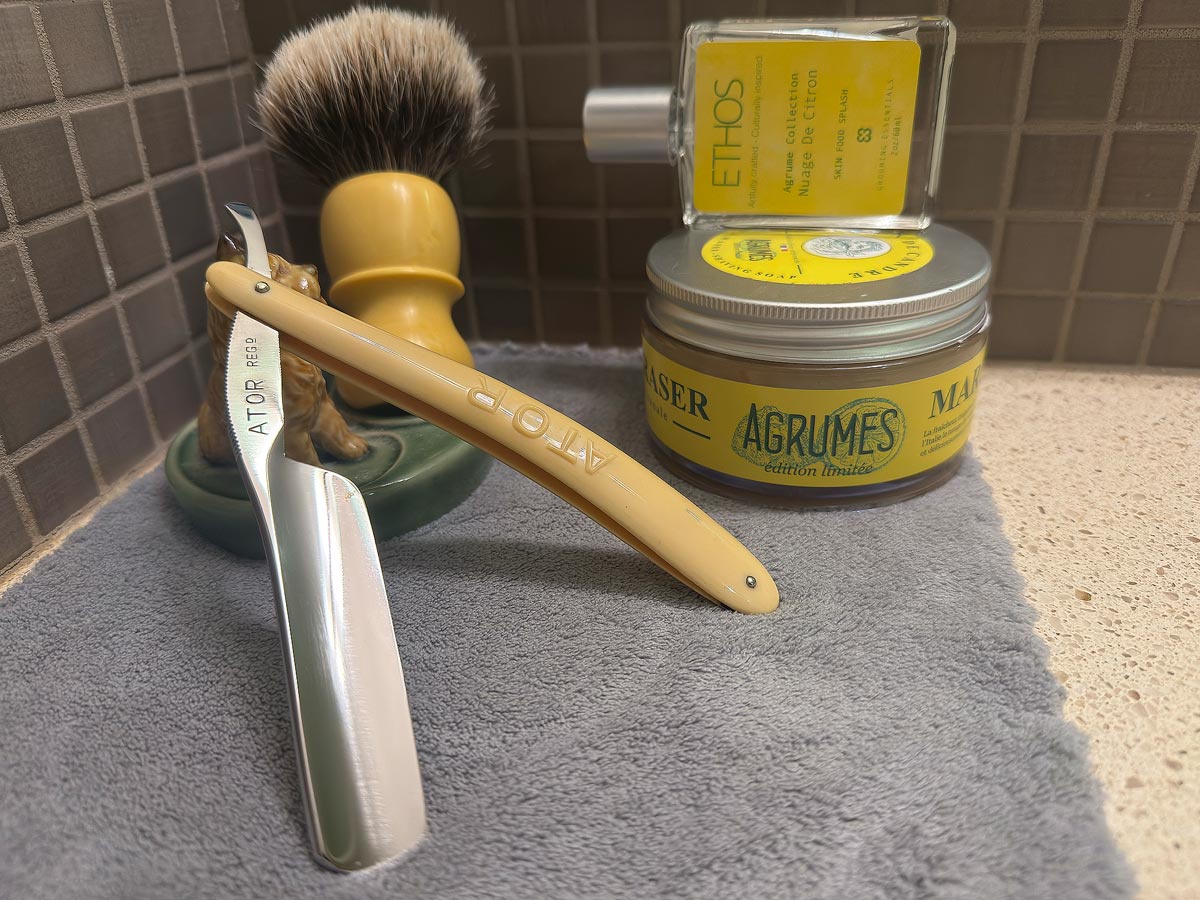 On a gray microfiber towel a straight razor with blade open about 45 degrees leans next to a bottle of aftershave and a jar of shave soap. A shave brush rests bristles-up in a green Wade ceramic pipe rest that features a brown Cairn Terrier.