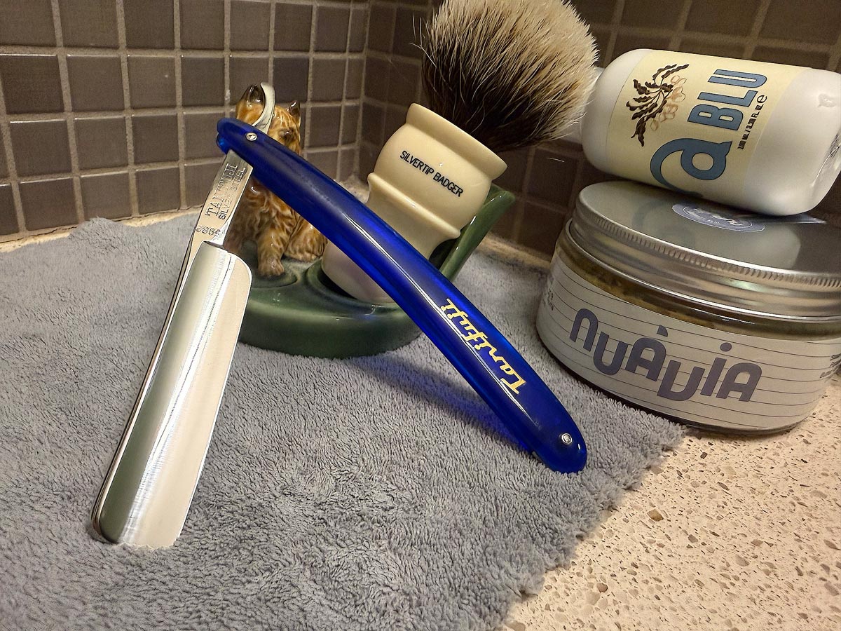 On a gray microfiber towel a straight razor with blade open about 45 degrees leans next to a bottle of aftershave and a jar of shave soap. A shave brush rests bristles-up in a green Wade ceramic pipe rest that features a brown Cairn Terrier.