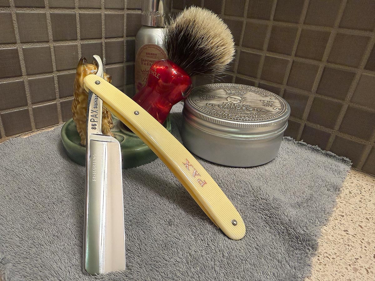 On a gray microfiber towel a straight razor with blade open about 45 degrees leans next to a bottle of aftershave and a jar of shave soap. A shave brush rests bristles-up in a green Wade ceramic pipe rest that features a brown Cairn Terrier.