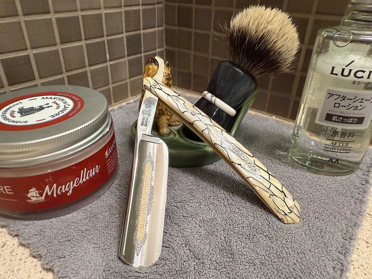 On a gray microfiber towel a straight razor with blade open about 45 degrees leans next to a bottle of aftershave and a jar of shave soap. A shave brush rests bristles-up in a green Wade ceramic pipe rest that features a brown Cairn Terrier.