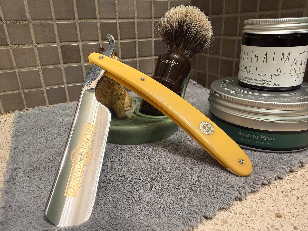 On a gray microfiber towel a straight razor with blade open about 45 degrees leans next to a bottle of aftershave and a jar of shave soap. A shave brush rests bristles-up in a green Wade ceramic pipe rest that features a brown Cairn Terrier.