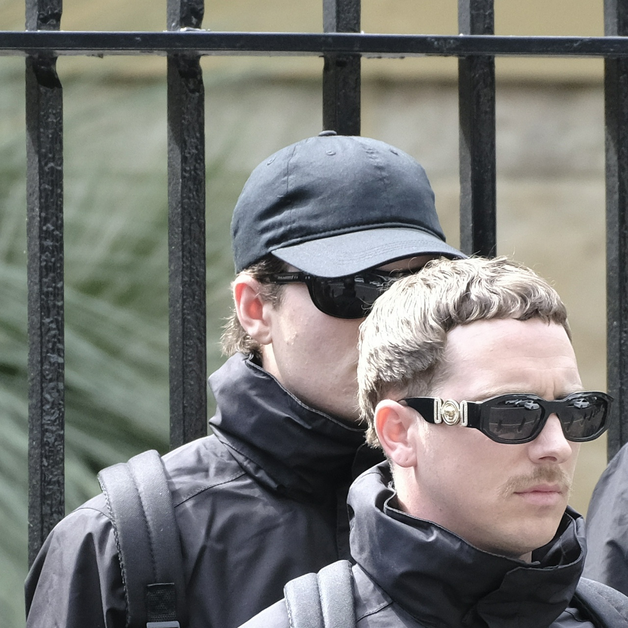A pimply man with brown hair, sunglasses and a cap hides behind his shorter gaudy companion