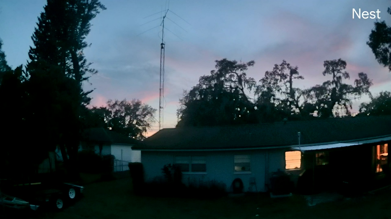 A backyard view just after sunset showing a Tempest Weather Station mounted high above a house, silhouetted against a blue, pink, and purple evening sky.