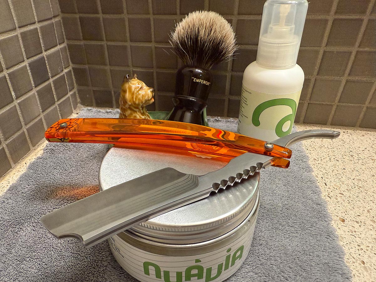 On a gray microfiber towel a straight razor with blade open about 45 degrees leans next to a bottle of aftershave and a jar of shave soap. A shave brush rests bristles-up in a green Wade ceramic pipe rest that features a brown Cairn Terrier.