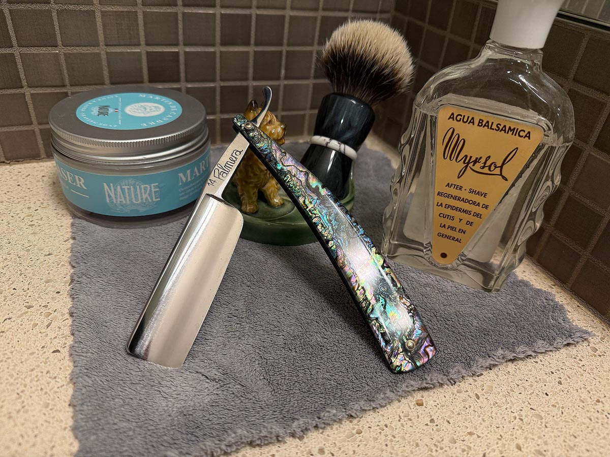 On a gray microfiber towel a straight razor with blade open about 45 degrees leans next to a bottle of aftershave and a jar of shave soap. A shave brush rests bristles-up in a green Wade ceramic pipe rest that features a brown Cairn Terrier.