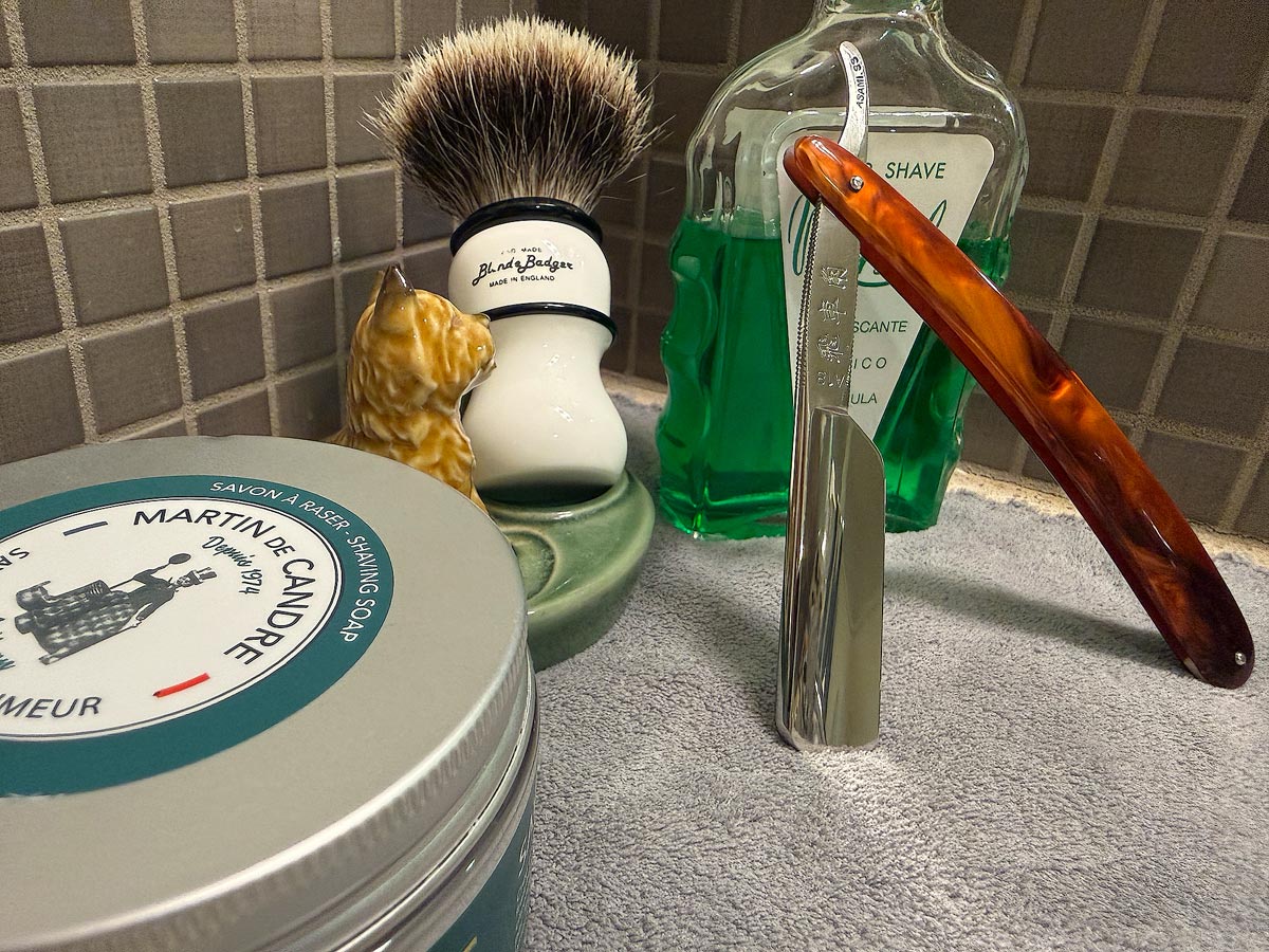 On a gray microfiber towel a straight razor with blade open about 45 degrees leans next to a bottle of aftershave and a jar of shave soap. A shave brush rests bristles-up in a green Wade ceramic pipe rest that features a brown Cairn Terrier.