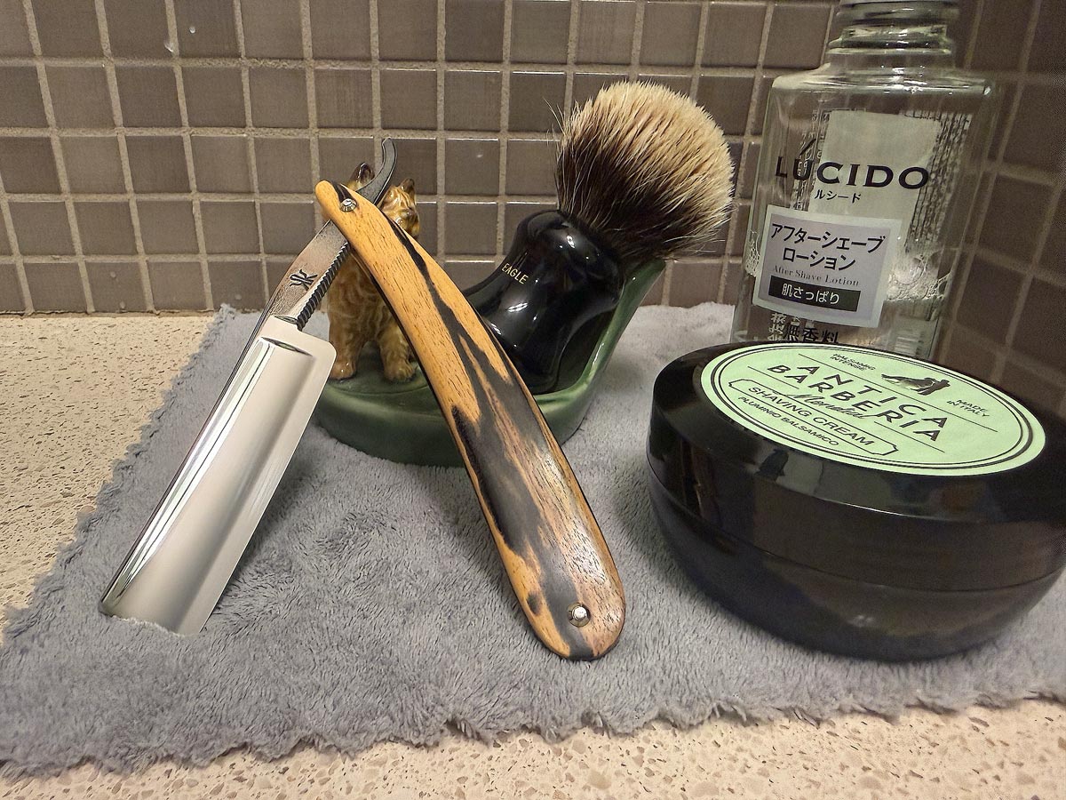 On a gray microfiber towel a straight razor with blade open about 45 degrees leans next to a bottle of aftershave and a jar of shave soap. A shave brush rests bristles-up in a green Wade ceramic pipe rest that features a brown Cairn Terrier.