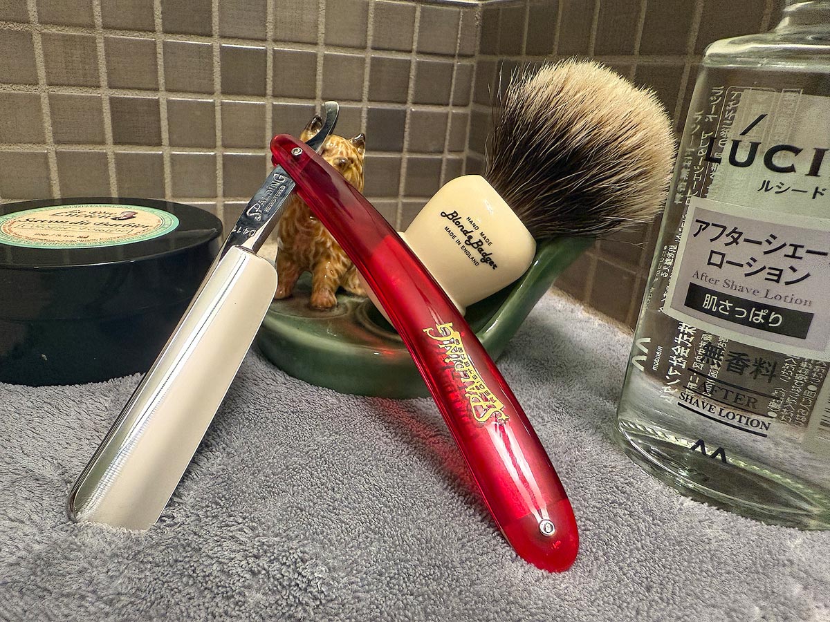 On a gray microfiber towel a straight razor with blade open about 45 degrees leans next to a bottle of aftershave and a jar of shave soap. A shave brush rests bristles-up in a green Wade ceramic pipe rest that features a brown Cairn Terrier.