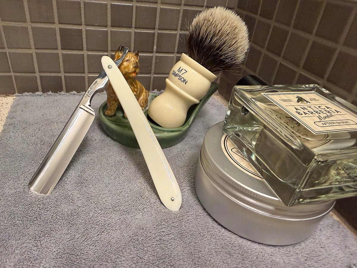 On a gray microfiber towel a straight razor with blade open about 45 degrees leans next to a bottle of aftershave and a jar of shave soap. A shave brush rests bristles-up in a green Wade ceramic pipe rest that features a brown Cairn Terrier.