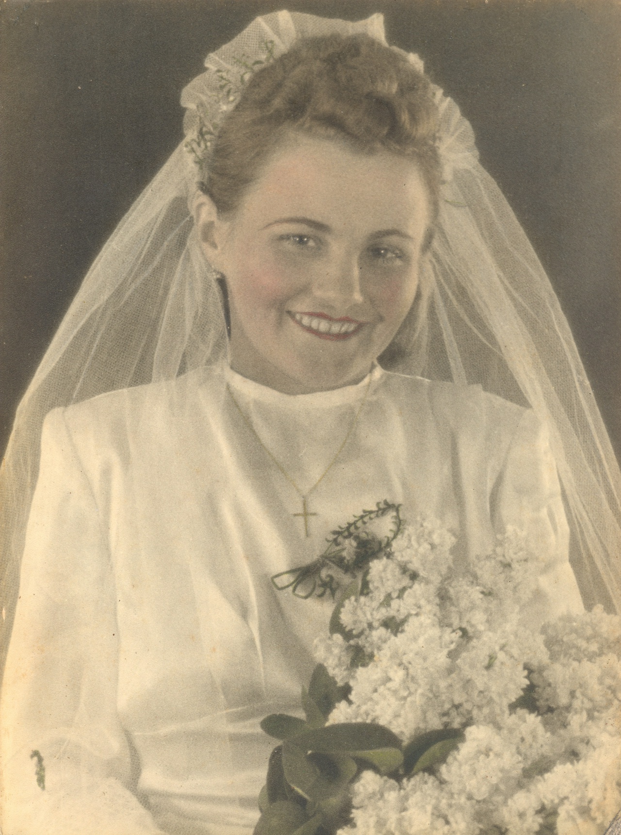 A vintage portrait of a smiling bride in her wedding attire. She wears a white gown with a high collar and a delicate cross necklace. Her hair is styled in elegant curls topped with a floral headpiece and a flowing veil. She holds a large bouquet of white flowers, adding a classic touch to her joyful expression.