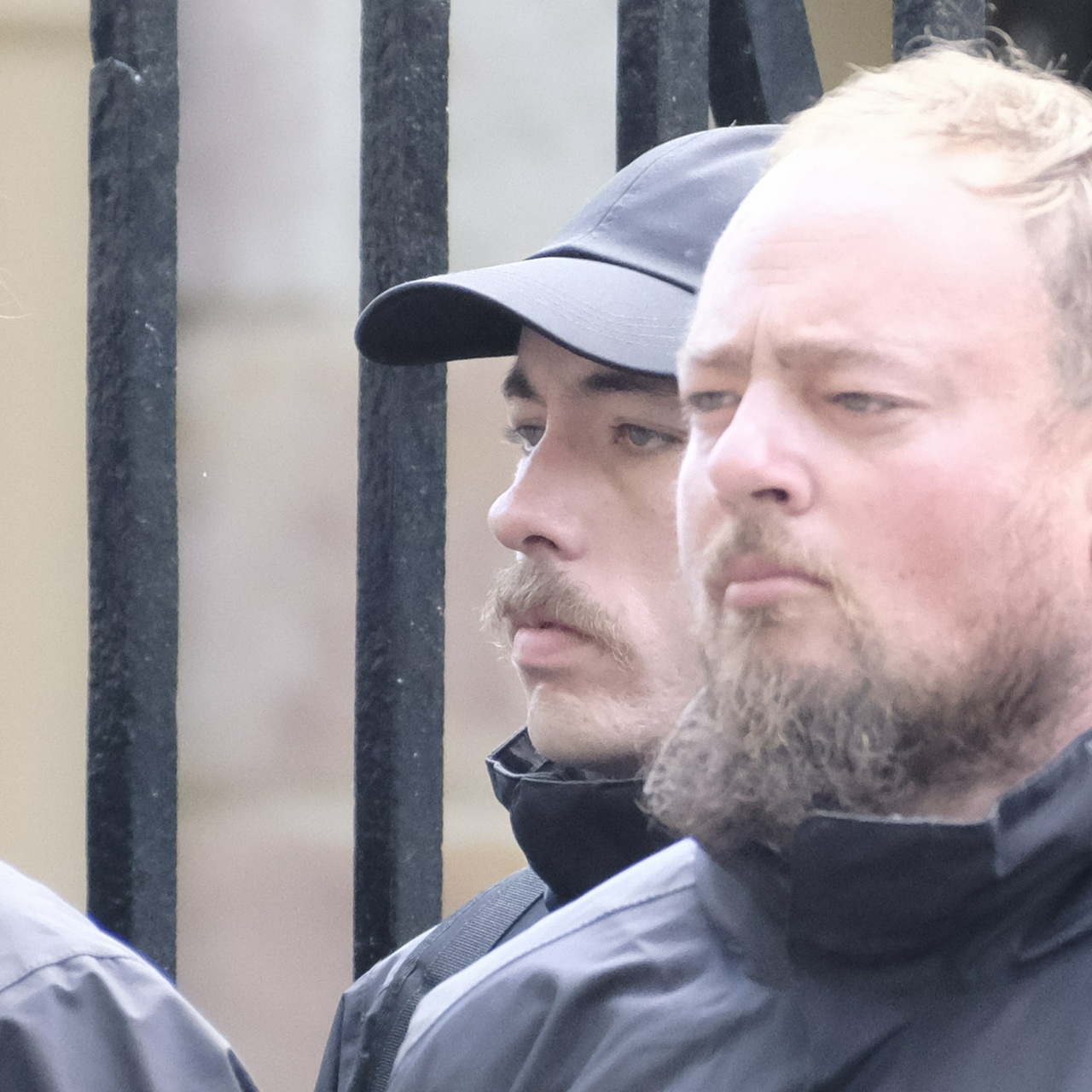 The same man peeking out from behind someone, showing his moustache almost jutting out as far as his nose