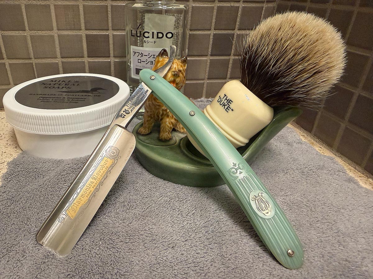 On a gray microfiber towel a straight razor with blade open about 45 degrees leans next to a bottle of aftershave and a jar of shave soap. A shave brush rests bristles-up in a green Wade ceramic pipe rest that features a brown Cairn Terrier.
