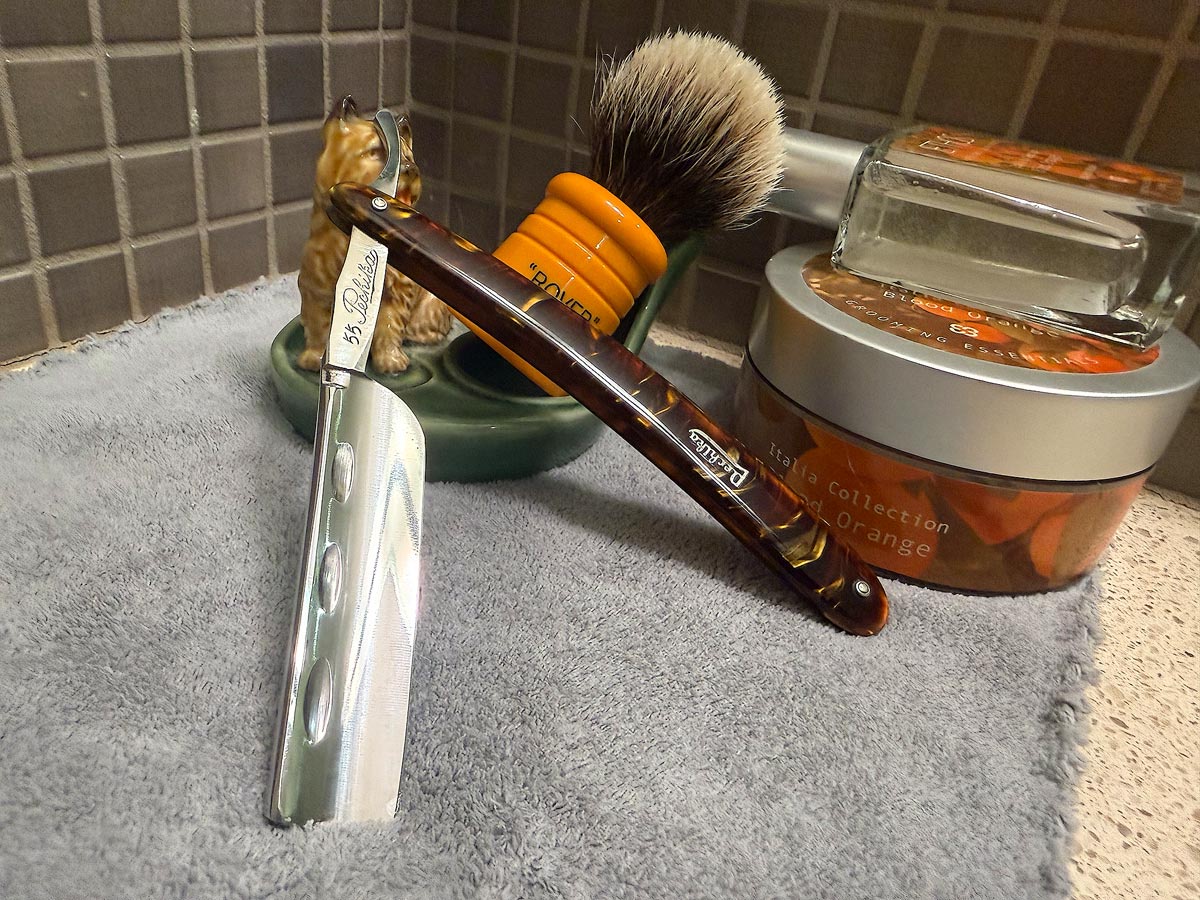 On a gray microfiber towel a straight razor with blade open about 45 degrees leans next to a bottle of aftershave and a jar of shave soap. A shave brush rests bristles-up in a green Wade ceramic pipe rest that features a brown Cairn Terrier.