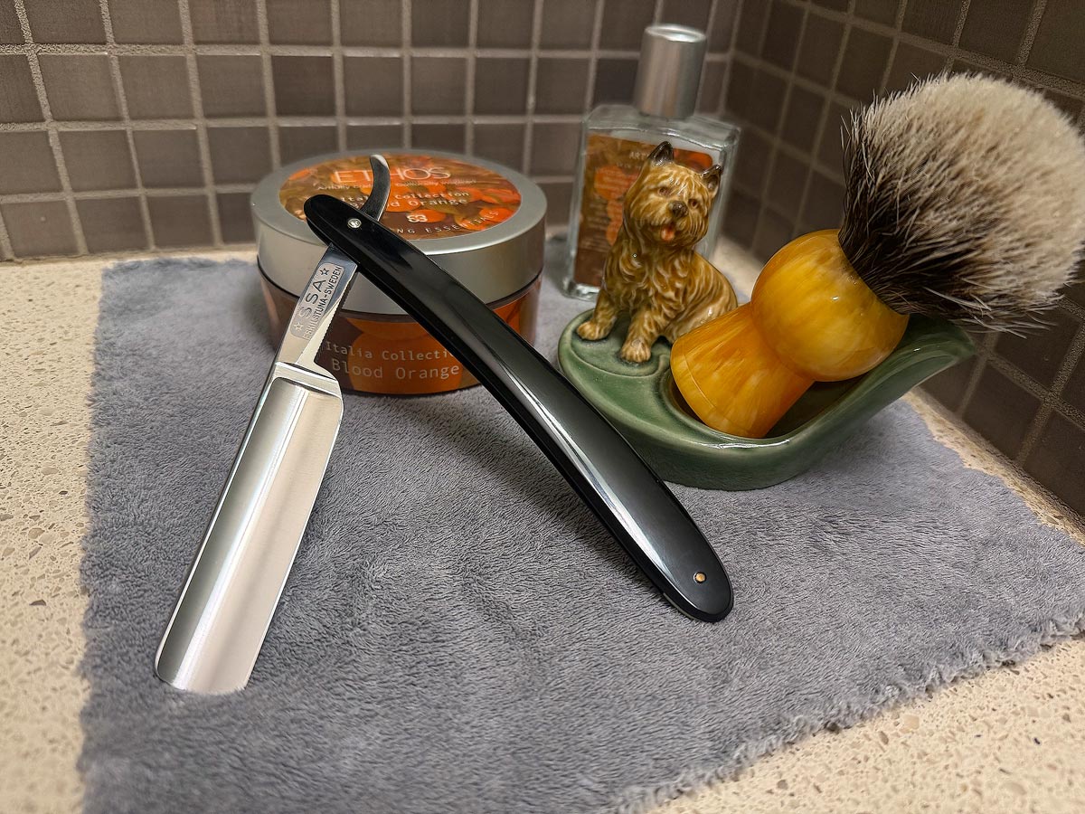 On a gray microfiber towel a straight razor with blade open about 45 degrees leans next to a bottle of aftershave and a jar of shave soap. A shave brush rests bristles-up in a green Wade ceramic pipe rest that features a brown Cairn Terrier.