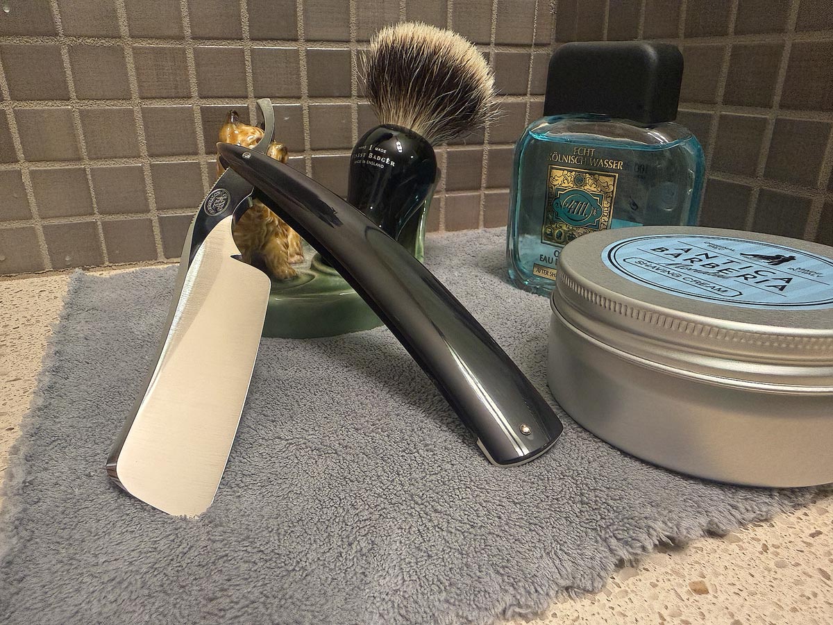 On a gray microfiber towel a straight razor with blade open about 45 degrees leans next to a bottle of aftershave and a jar of shave soap. A shave brush rests bristles-up in a green Wade ceramic pipe rest that features a brown Cairn Terrier.