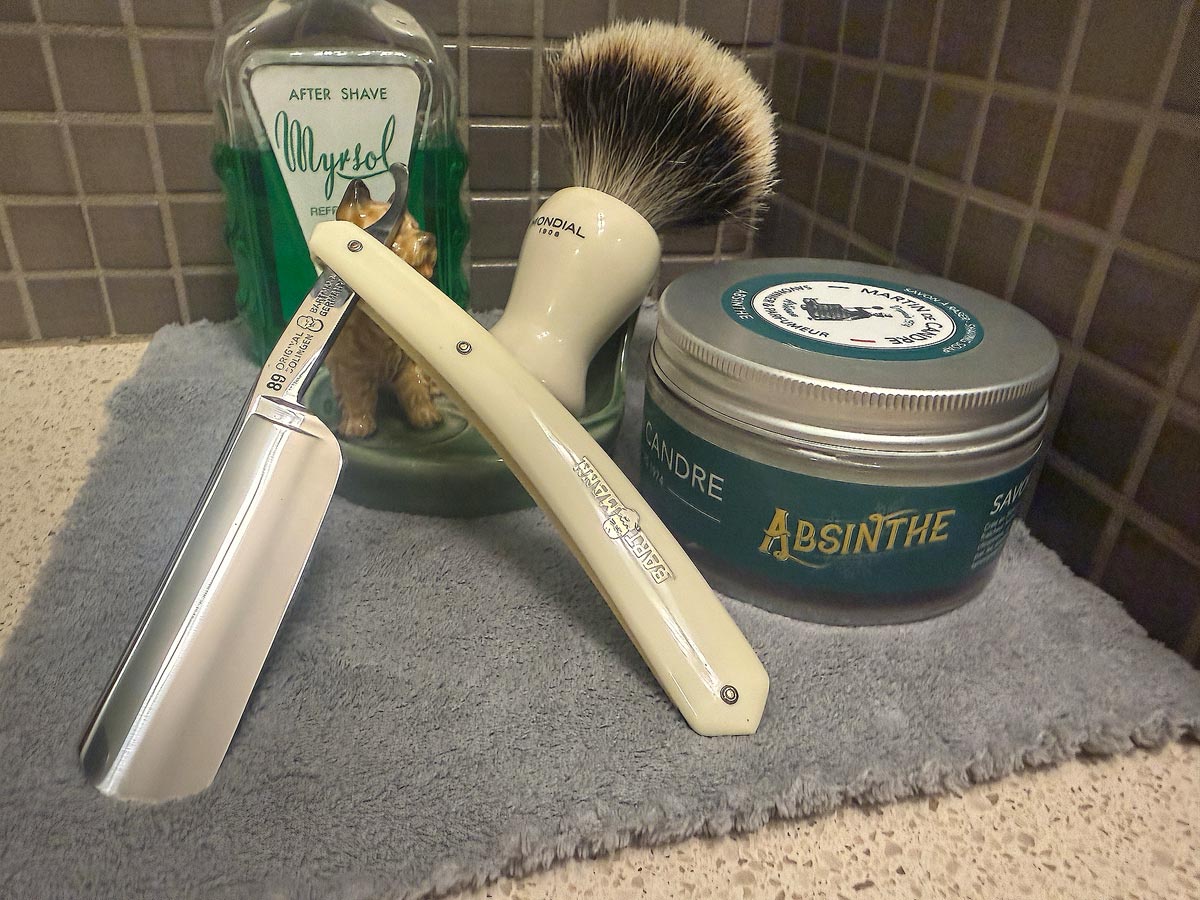 On a gray microfiber towel a straight razor with blade open about 45 degrees leans next to a bottle of aftershave and a jar of shave soap. A shave brush rests bristles-up in a green Wade ceramic pipe rest that features a brown Cairn Terrier.
