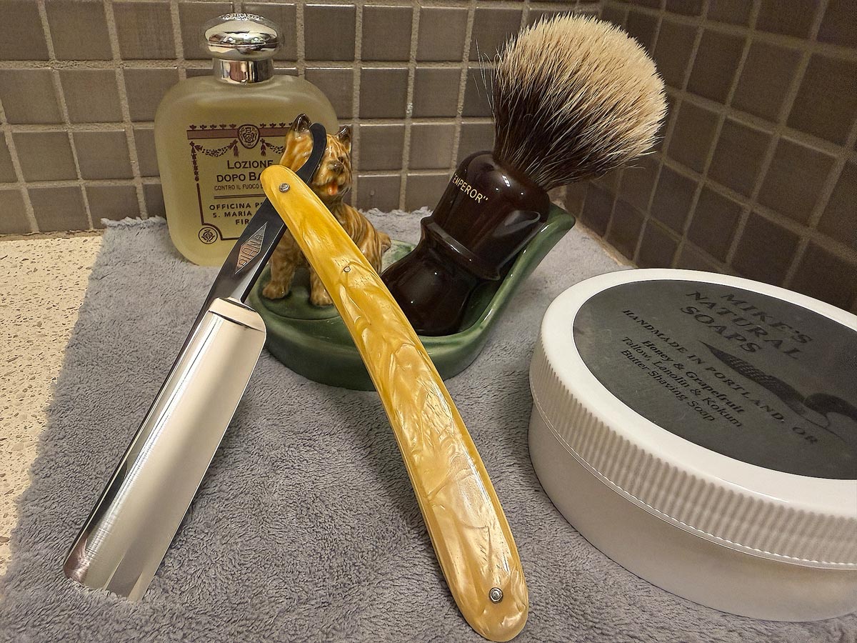 On a gray microfiber towel a straight razor with blade open about 45 degrees leans next to a bottle of aftershave and a jar of shave soap. A shave brush rests bristles-up in a green Wade ceramic pipe rest that features a brown Cairn Terrier.