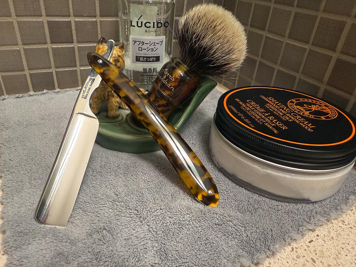 On a gray microfiber towel a straight razor with blade open about 45 degrees leans next to a bottle of aftershave and a jar of shave soap. A shave brush rests bristles-up in a green Wade ceramic pipe rest that features a brown Cairn Terrier.