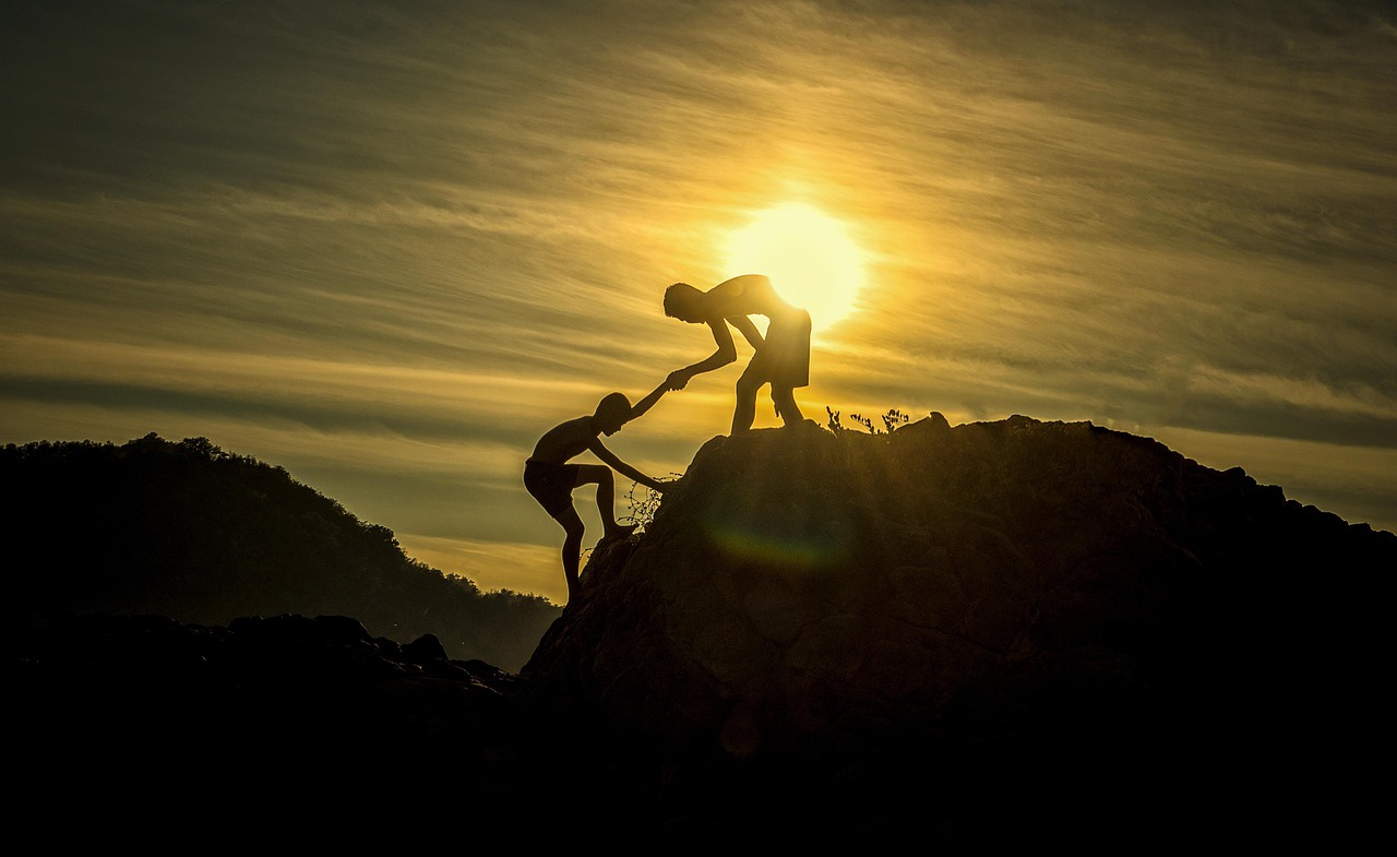 Two people silhouetted by the sun climb a steep hill top. The one already on the summit holds the hand of the one still rising.