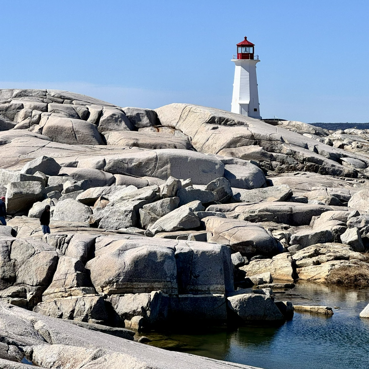 Peggy's Cove lighthouse, white with a red top, rises above a landscape of large smooth granite boulders on the Atlantic coast. A small pool of water sits in the foreground between the rocks. Two small figures are visible climbing the rocks in the middle distance. Clear blue sky.