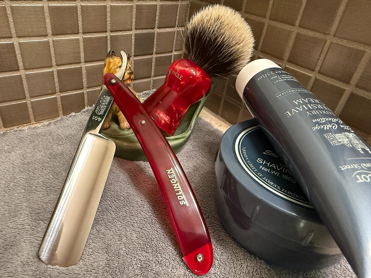 On a gray microfiber towel a straight razor with blade open about 45 degrees leans next to a bottle of aftershave and a jar of shave soap. A shave brush rests bristles-up in a green Wade ceramic pipe rest that features a brown Cairn Terrier.