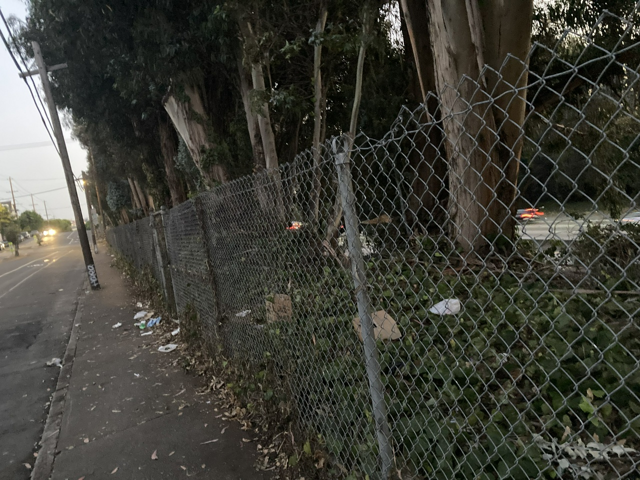 a trash covered stretch of sidewalk next to a chain link fence with trees and a hillside and a busy highway below