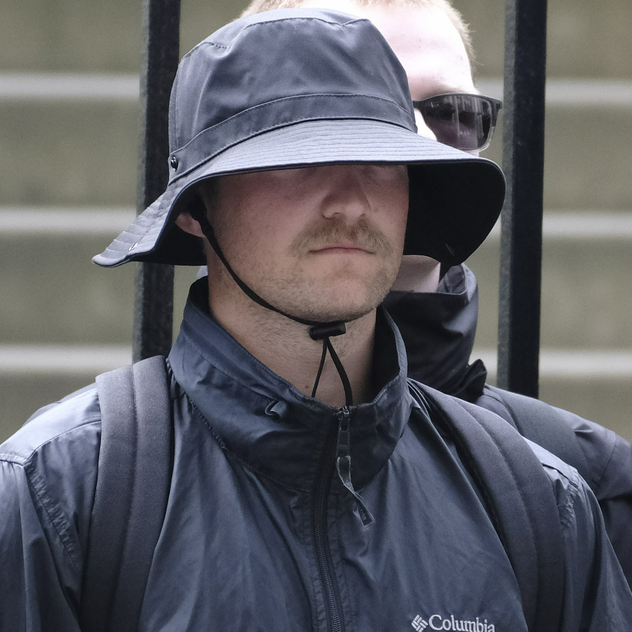 An unshaven blonde man with a moustache and a bucket hat fastened so tight under his chin that it's digging in