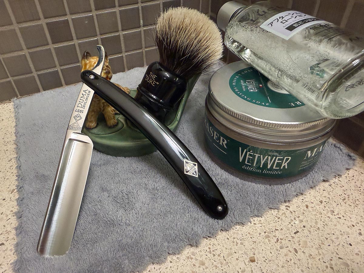 On a gray microfiber towel a straight razor with blade open about 45 degrees leans next to a bottle of aftershave and a jar of shave soap. A shave brush rests bristles-up in a green Wade ceramic pipe rest that features a brown Cairn Terrier.