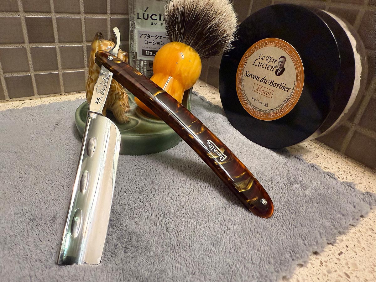 On a gray microfiber towel a straight razor with blade open about 45 degrees leans next to a bottle of aftershave and a jar of shave soap. A shave brush rests bristles-up in a green Wade ceramic pipe rest that features a brown Cairn Terrier.