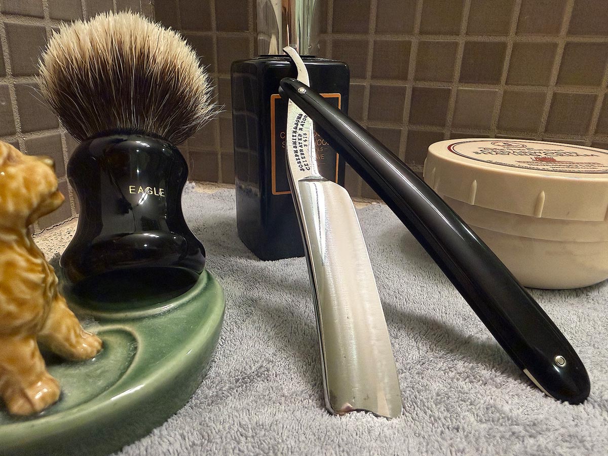On a gray microfiber towel a straight razor with blade open about 45 degrees leans next to a bottle of aftershave and a jar of shave soap. A shave brush rests bristles-up in a green Wade ceramic pipe rest that features a brown Cairn Terrier.