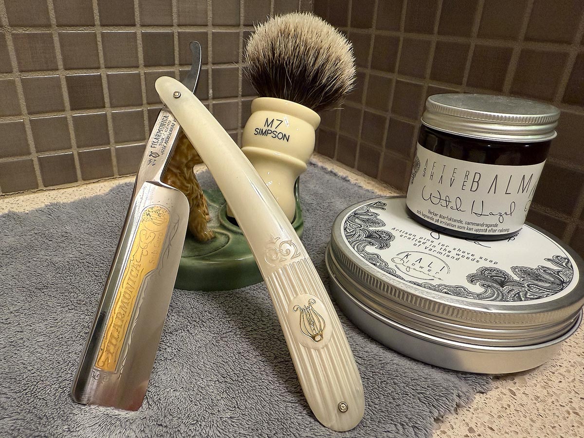 On a gray microfiber towel a straight razor with blade open about 45 degrees leans next to a bottle of aftershave and a jar of shave soap. A shave brush rests bristles-up in a green Wade ceramic pipe rest that features a brown Cairn Terrier.