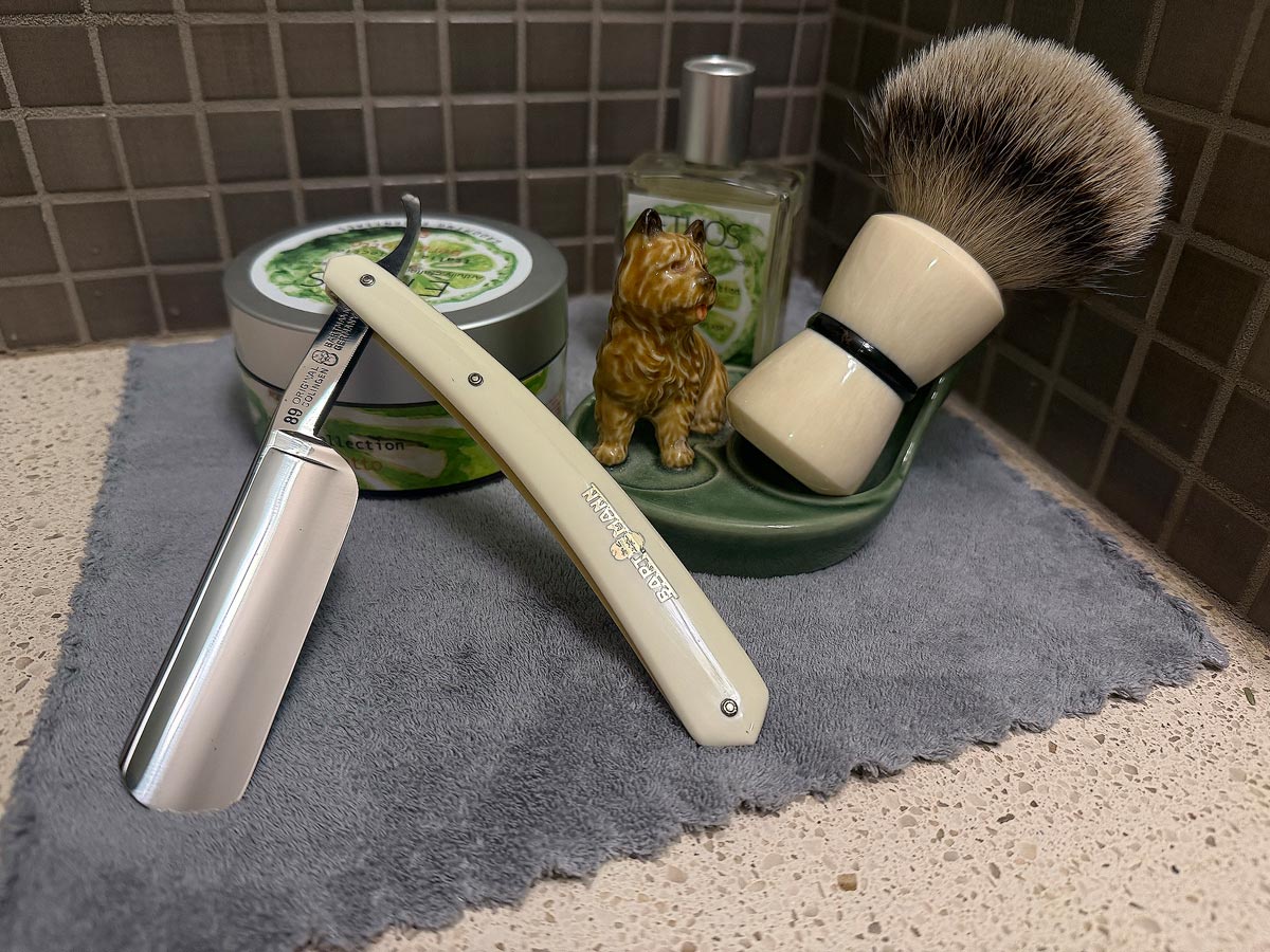 On a gray microfiber towel a straight razor with blade open about 45 degrees leans next to a bottle of aftershave and a jar of shave soap. A shave brush rests bristles-up in a green Wade ceramic pipe rest that features a brown Cairn Terrier.
