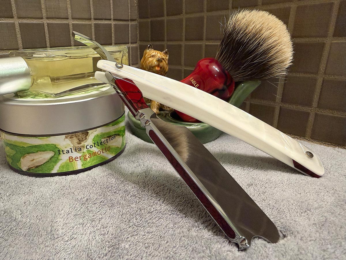 On a gray microfiber towel a straight razor with blade open about 45 degrees leans next to a bottle of aftershave and a jar of shave soap. A shave brush rests bristles-up in a green Wade ceramic pipe rest that features a brown Cairn Terrier.