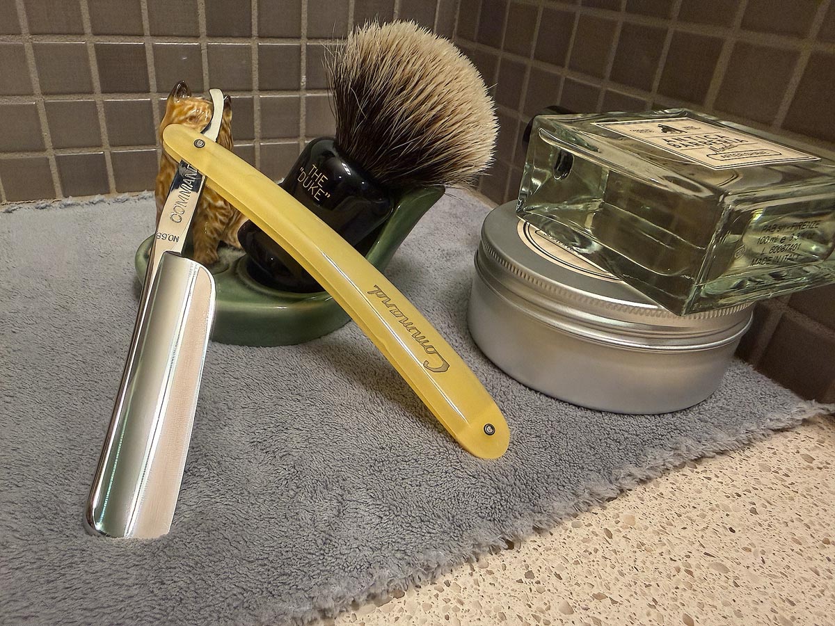 On a gray microfiber towel a straight razor with blade open about 45 degrees leans next to a bottle of aftershave and a jar of shave soap. A shave brush rests bristles-up in a green Wade ceramic pipe rest that features a brown Cairn Terrier.