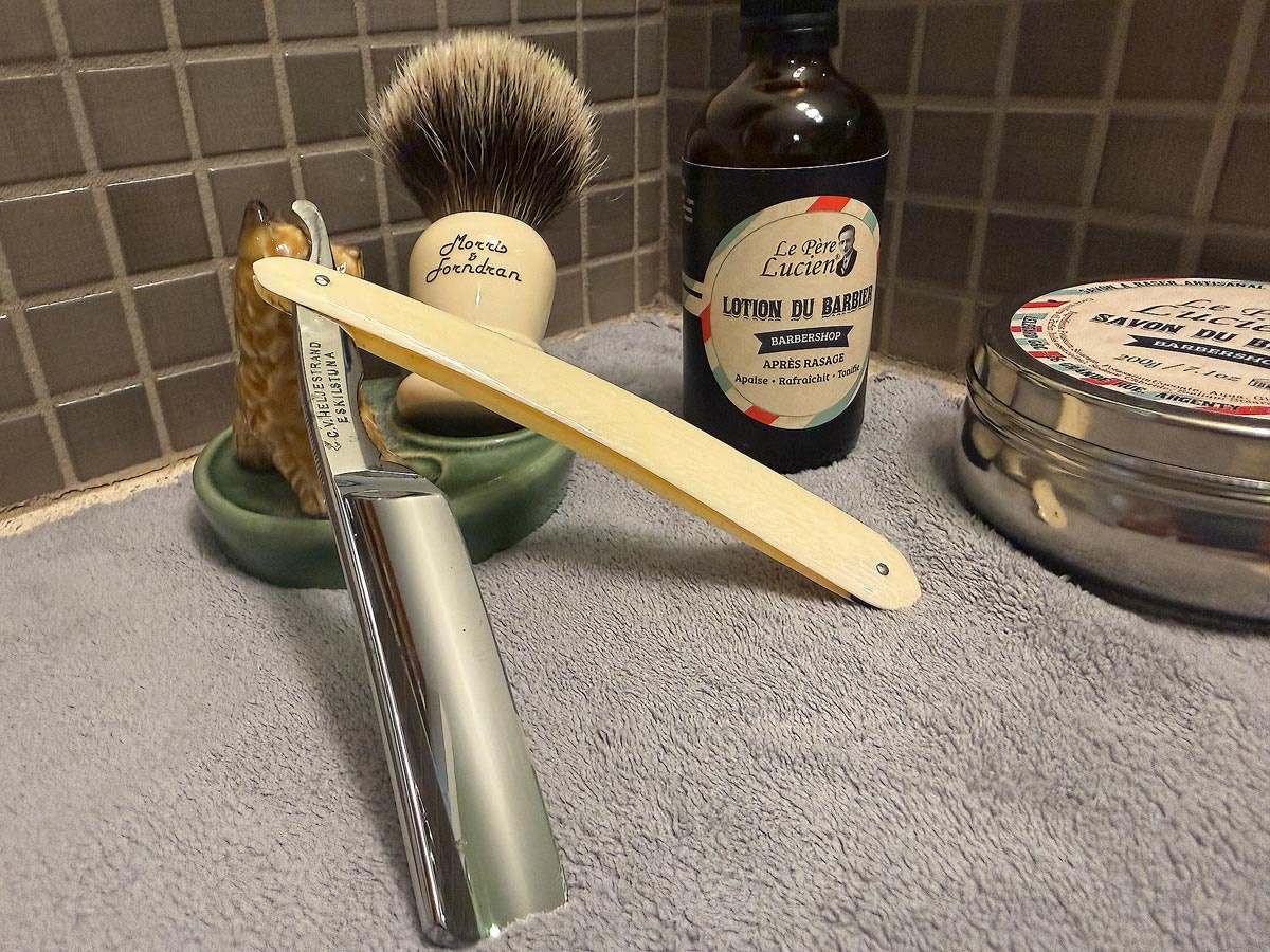 On a gray microfiber towel a straight razor with blade open about 45 degrees leans next to a bottle of aftershave and a jar of shave soap. A shave brush rests bristles-up in a green Wade ceramic pipe rest that features a brown Cairn Terrier.