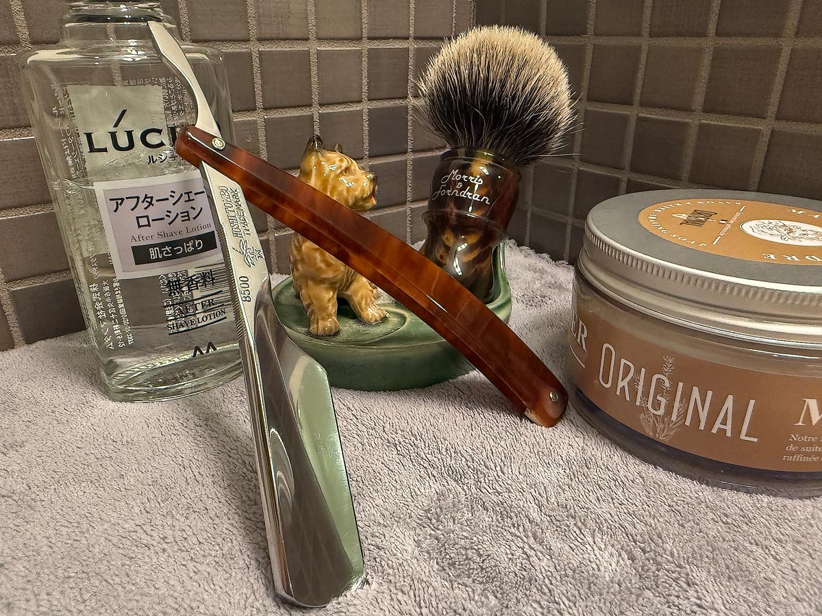 On a gray microfiber towel a straight razor with blade open about 45 degrees leans next to a bottle of aftershave and a jar of shave soap. A shave brush rests bristles-up in a green Wade ceramic pipe rest that features a brown Cairn Terrier.