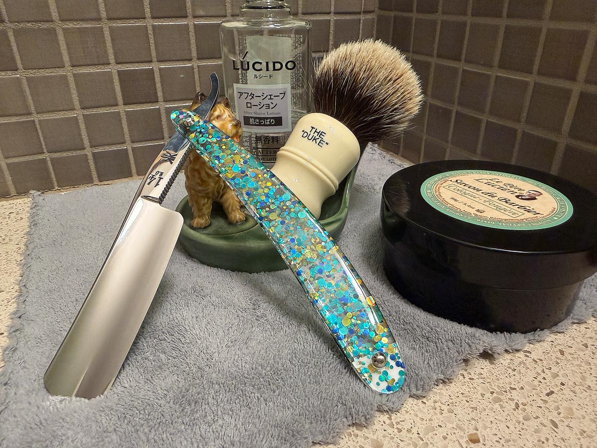 On a gray microfiber towel a straight razor with blade open about 45 degrees leans next to a bottle of aftershave and a jar of shave soap. A shave brush rests bristles-up in a green Wade ceramic pipe rest that features a brown Cairn Terrier.