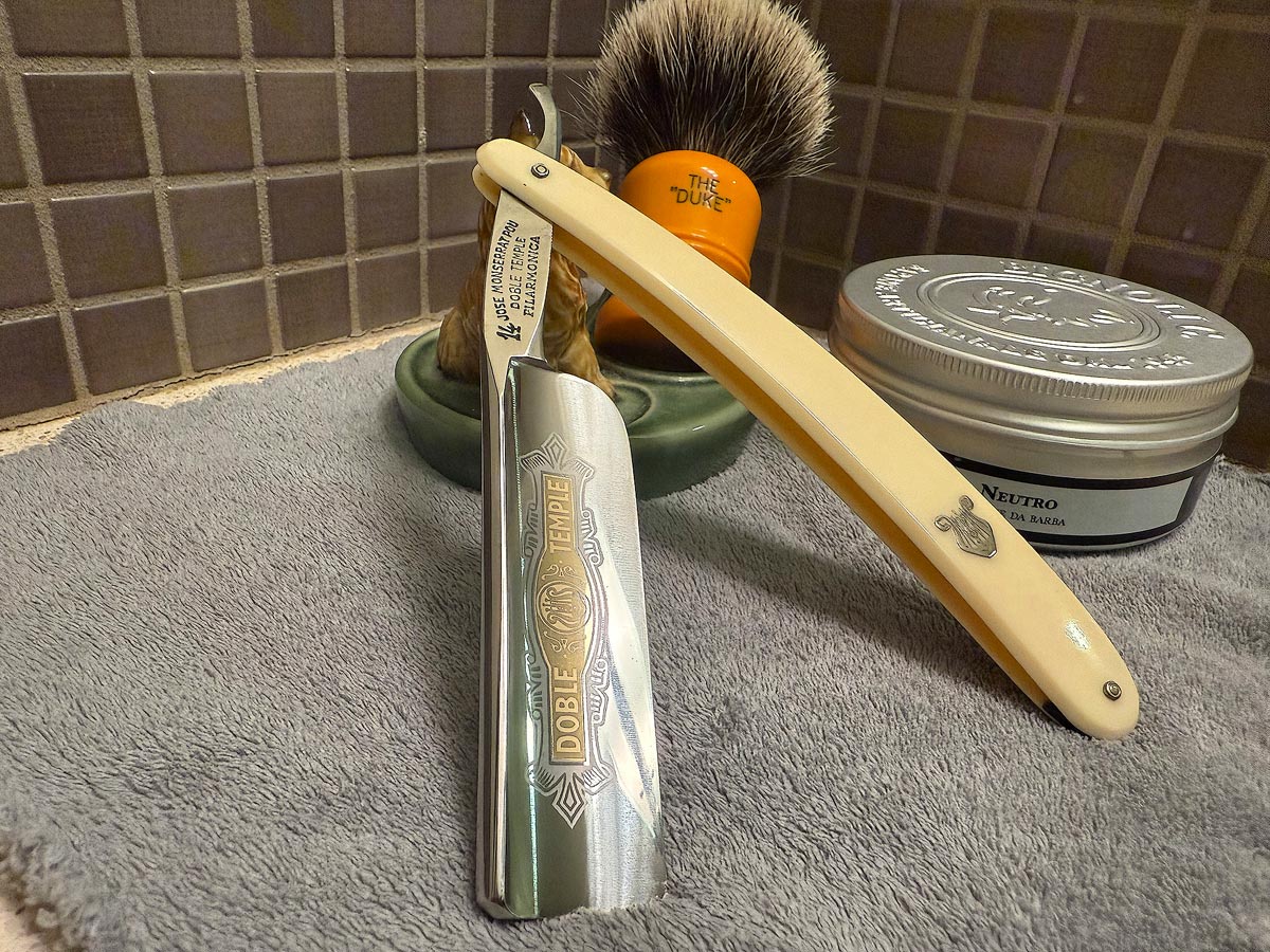 On a gray microfiber towel a straight razor with blade open about 45 degrees leans next to a bottle of aftershave and a jar of shave soap. A shave brush rests bristles-up in a green Wade ceramic pipe rest that features a brown Cairn Terrier.