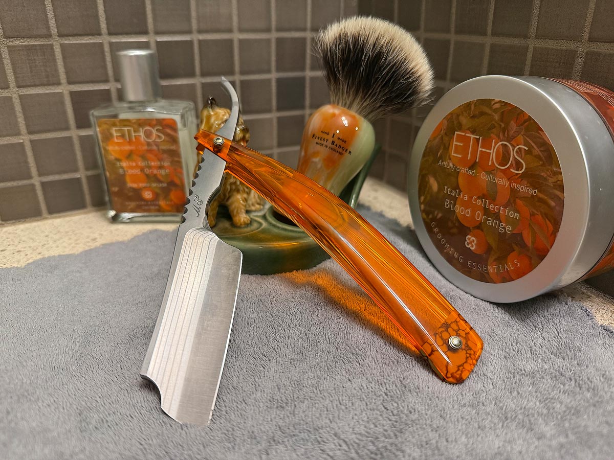 On a gray microfiber towel a straight razor with blade open about 45 degrees leans next to a bottle of aftershave and a jar of shave soap. A shave brush rests bristles-up in a green Wade ceramic pipe rest that features a brown Cairn Terrier.