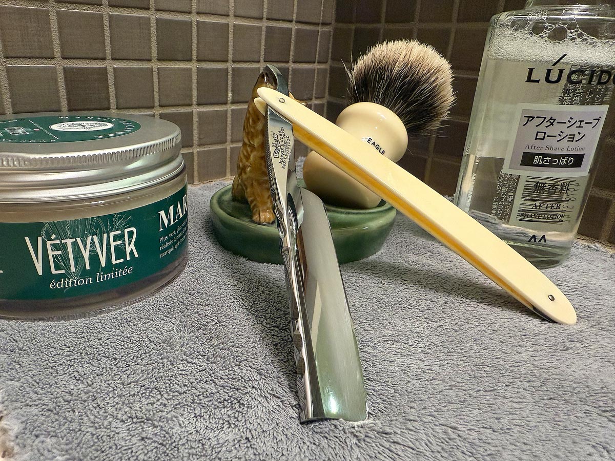 On a gray microfiber towel a straight razor with blade open about 45 degrees leans next to a bottle of aftershave and a jar of shave soap. A shave brush rests bristles-up in a green Wade ceramic pipe rest that features a brown Cairn Terrier.