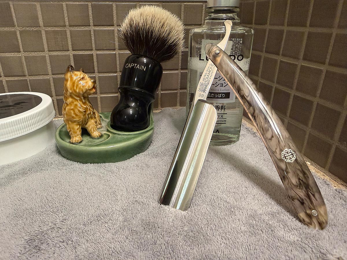 On a gray microfiber towel a straight razor with blade open about 45 degrees leans next to a bottle of aftershave and a jar of shave soap. A shave brush rests bristles-up in a green Wade ceramic pipe rest that features a brown Cairn Terrier.