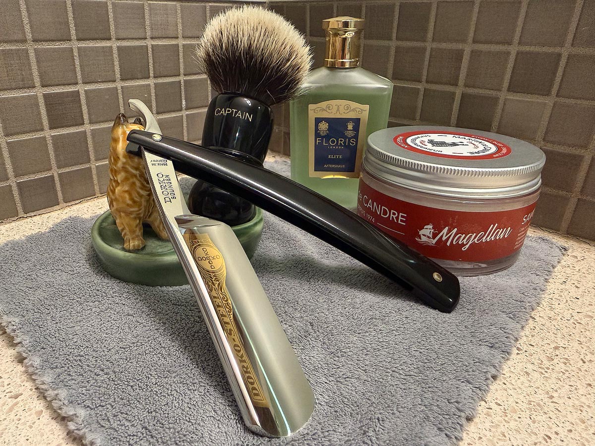 On a gray microfiber towel a straight razor with blade open about 45 degrees leans next to a bottle of aftershave and a jar of shave soap. A shave brush rests bristles-up in a green Wade ceramic pipe rest that features a brown Cairn Terrier.