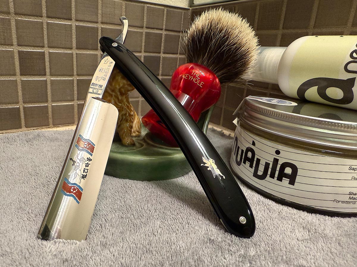 On a gray microfiber towel a straight razor with blade open about 45 degrees leans next to a bottle of aftershave and a jar of shave soap. A shave brush rests bristles-up in a green Wade ceramic pipe rest that features a brown Cairn Terrier.