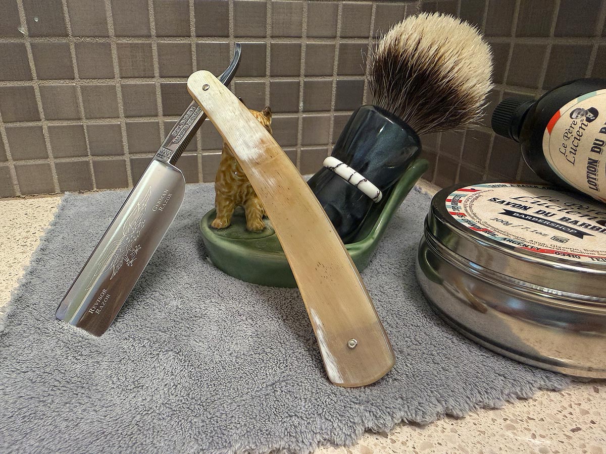 On a gray microfiber towel a straight razor with blade open about 45 degrees leans next to a bottle of aftershave and a jar of shave soap. A shave brush rests bristles-up in a green Wade ceramic pipe rest that features a brown Cairn Terrier.