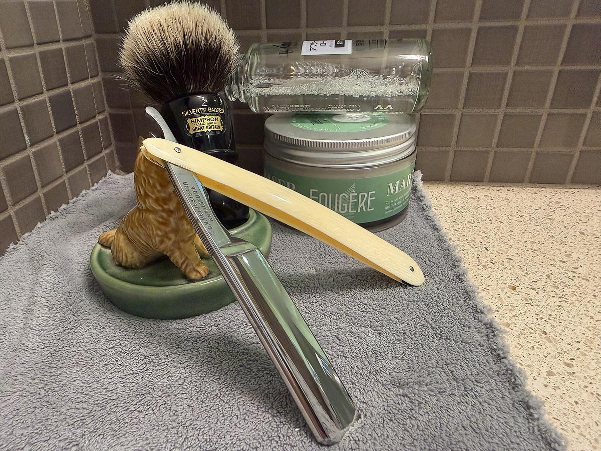 On a gray microfiber towel a straight razor with blade open about 45 degrees leans next to a bottle of aftershave and a jar of shave soap. A shave brush rests bristles-up in a green Wade ceramic pipe rest that features a brown Cairn Terrier.