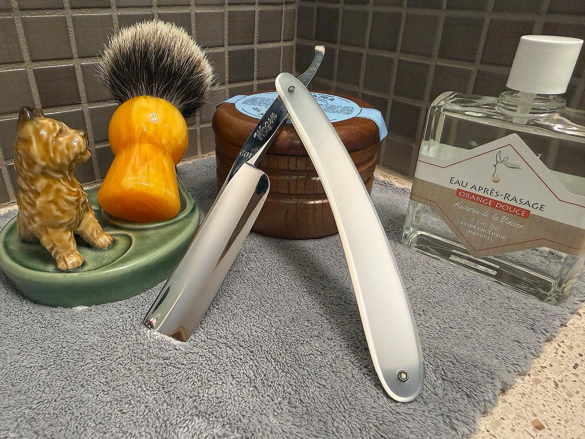 On a gray microfiber towel a straight razor with blade open about 45 degrees leans next to a bottle of aftershave and a jar of shave soap. A shave brush rests bristles-up in a green Wade ceramic pipe rest that features a brown Cairn Terrier.