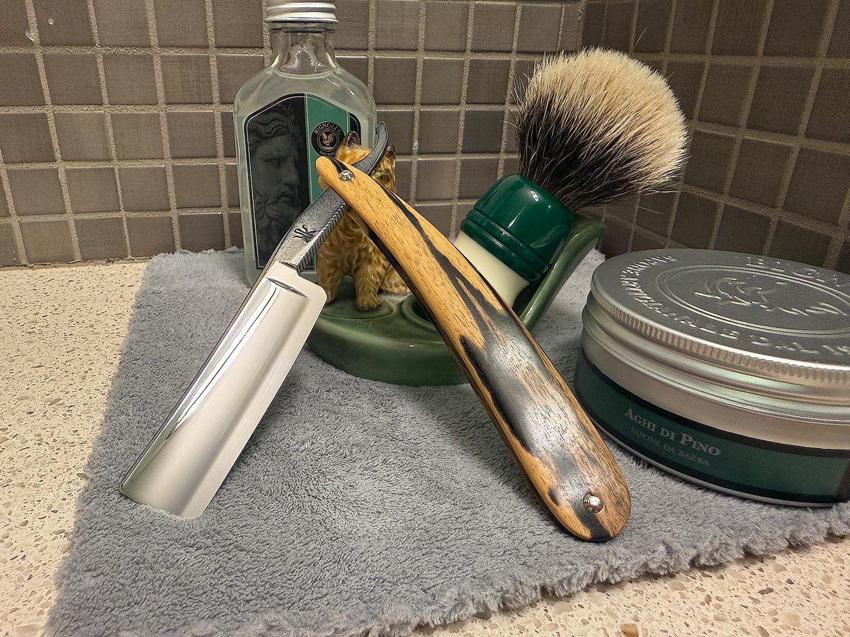 On a gray microfiber towel a straight razor with blade open about 45 degrees leans next to a bottle of aftershave and a jar of shave soap. A shave brush rests bristles-up in a green Wade ceramic pipe rest that features a brown Cairn Terrier.